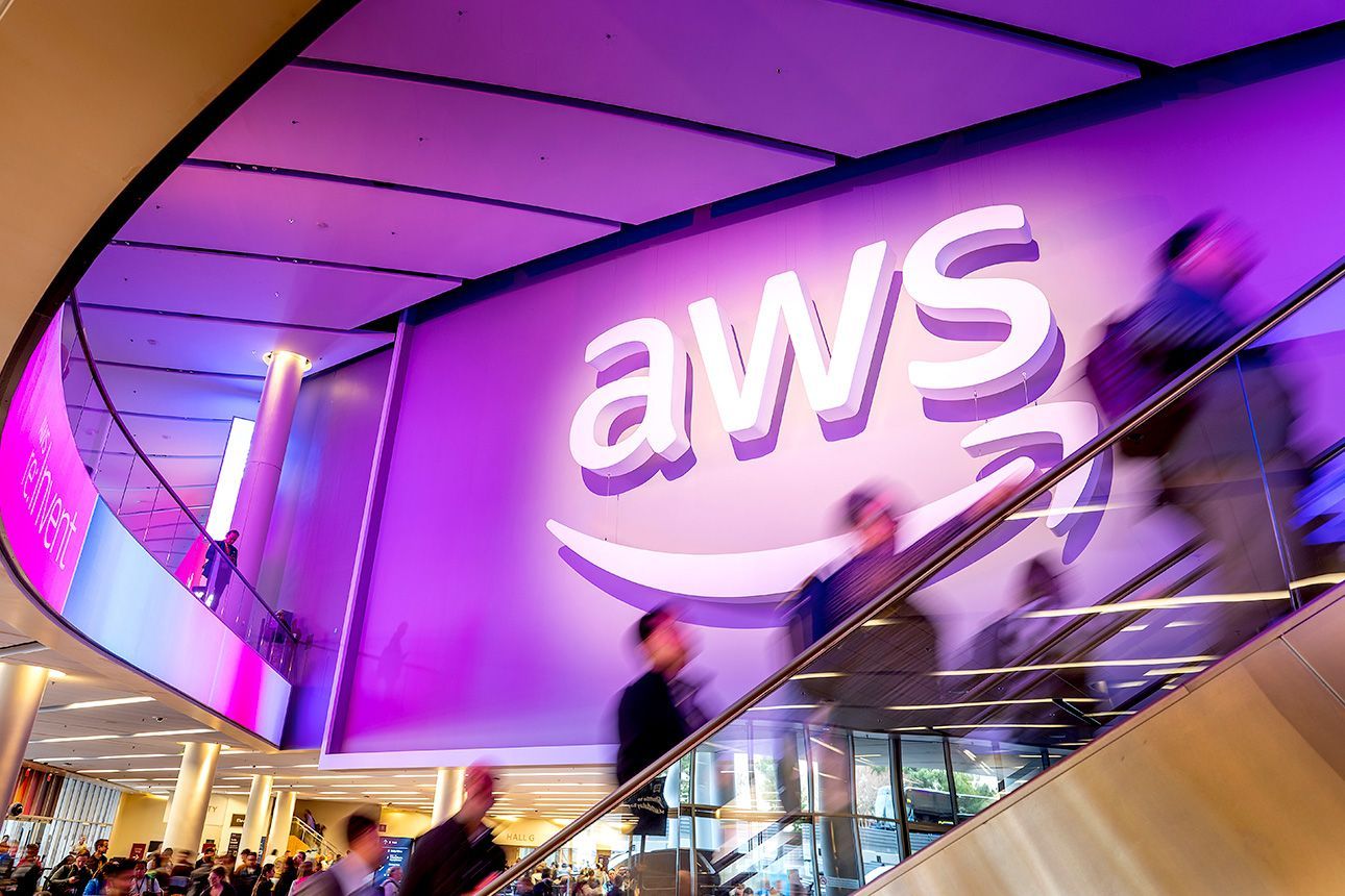 AWS logo displayed above a busy escalator at a purple-lit technology conference venue.