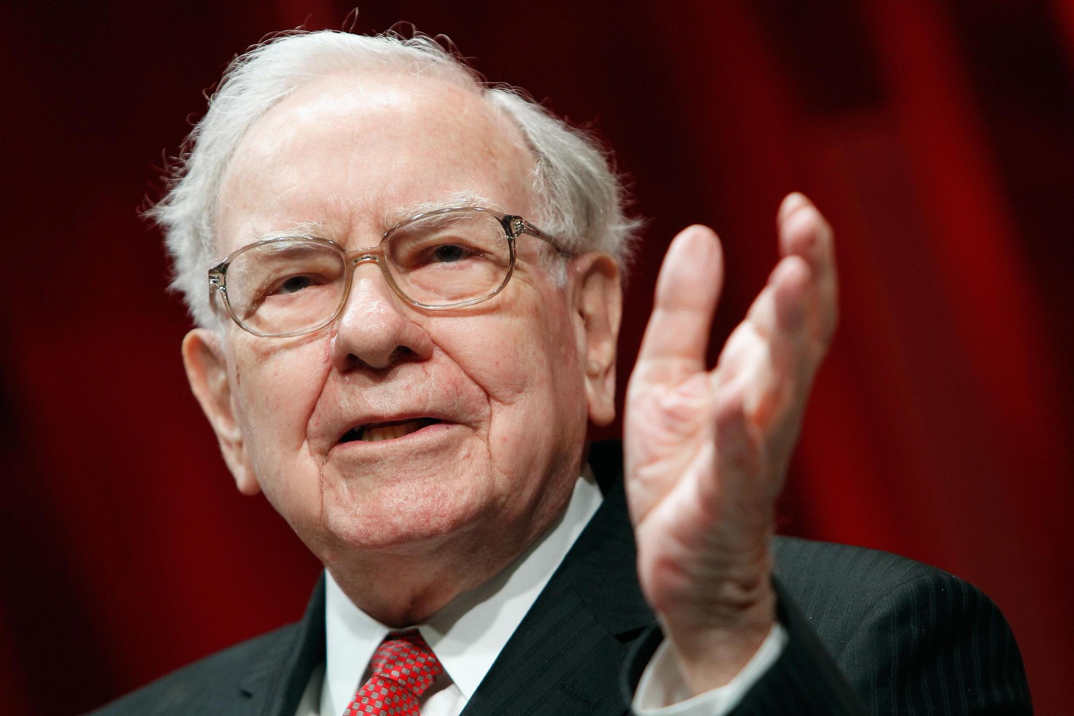 Warren Buffett gestures while speaking against a red background.